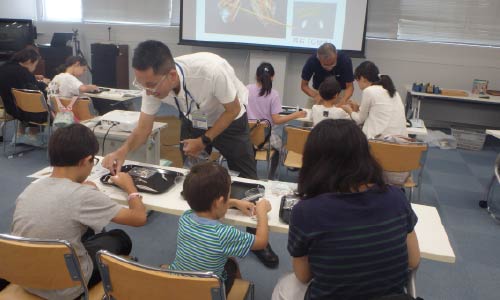 親子連れが講師から環境保全についてレクチャーを受けている。教室で椅子に座っている